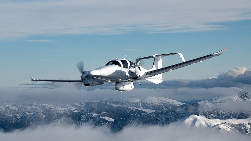 A sleek white aircraft soaring through a cloudy sky, with snow-capped mountains visible in the background.