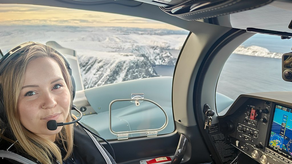 A female pilot smiles confidently in the cockpit, with picturesque snowy mountains and ocean visible in the background.