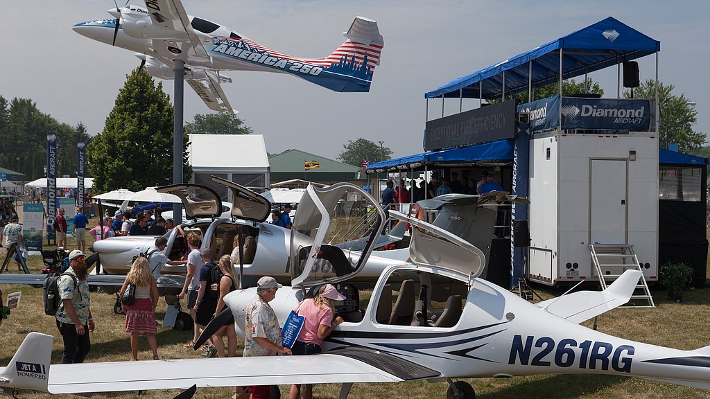 A busy airshow scene featuring several small aircraft, with people exploring and interacting around them under a clear sky.