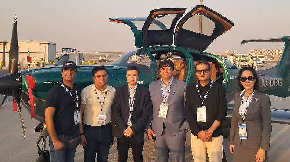 A group of six individuals stands beside a DA50 RG aircraft, showcasing a mix of formal and casual attire against a clear sky.
