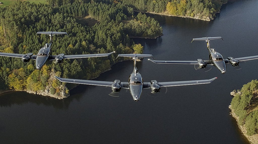 Three DA62 MPP aircraft fly in formation over a serene lake, surrounded by lush green forests and autumn-colored trees.