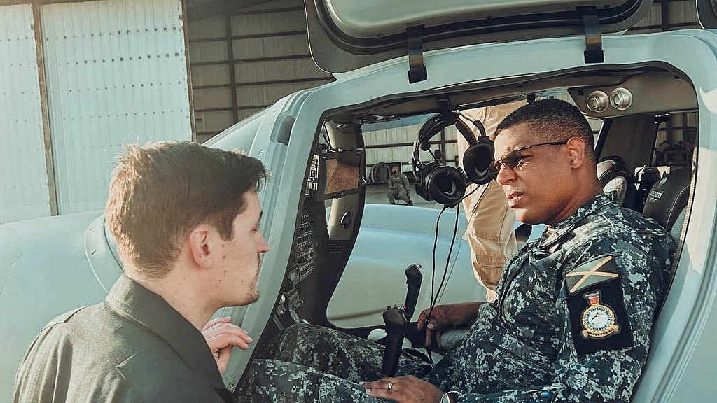 In a sunlit hangar, a soldier in camouflage talks to a colleague, who stands beside the open cockpit of a DA62 MPP.