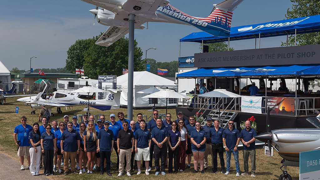 A large group of people stands together in front of a display featuring small aircraft and a promotional booth at an airshow.