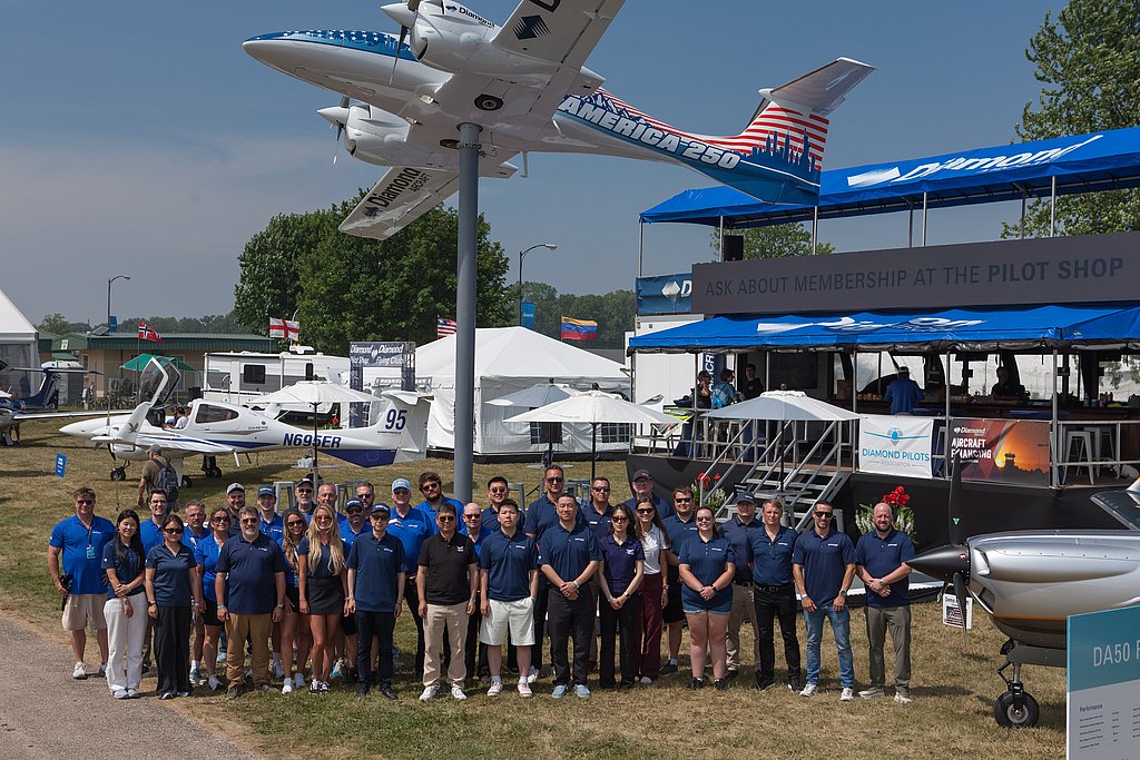 A large group of people stands together in front of a display featuring small aircraft and a promotional booth at an airshow.
