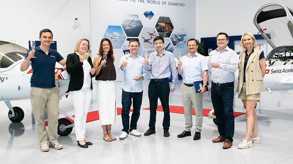 A group of eight people celebrate with drinks in hand, standing in front of a small aircraft in a bright, modern setting.