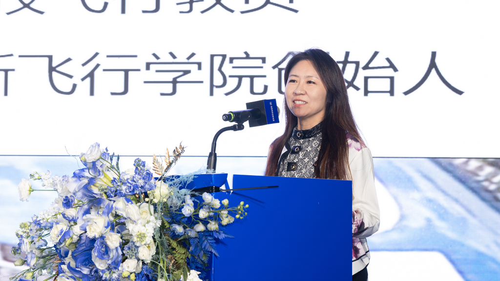 The speaker, dressed elegantly, engages the audience while presenting at a podium decorated with vibrant blue flowers.