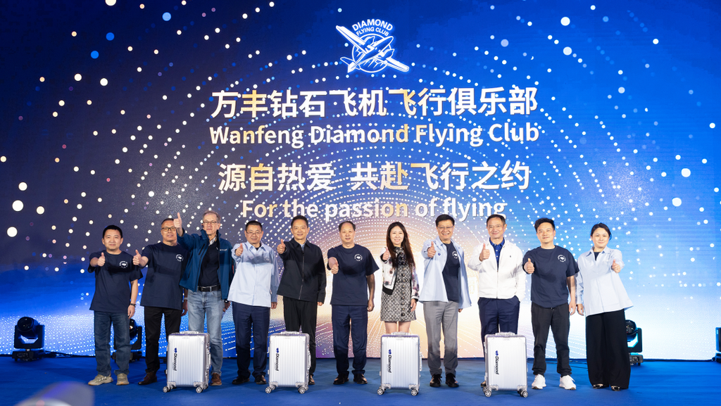 A group of eight individuals stands together, smiling and giving thumbs up, in front of a vibrant blue backdrop with logos.
