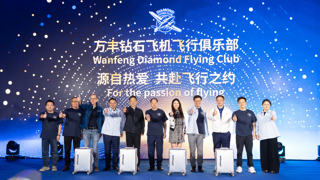A group of eight individuals stands together, smiling and giving thumbs up, in front of a vibrant blue backdrop with logos.