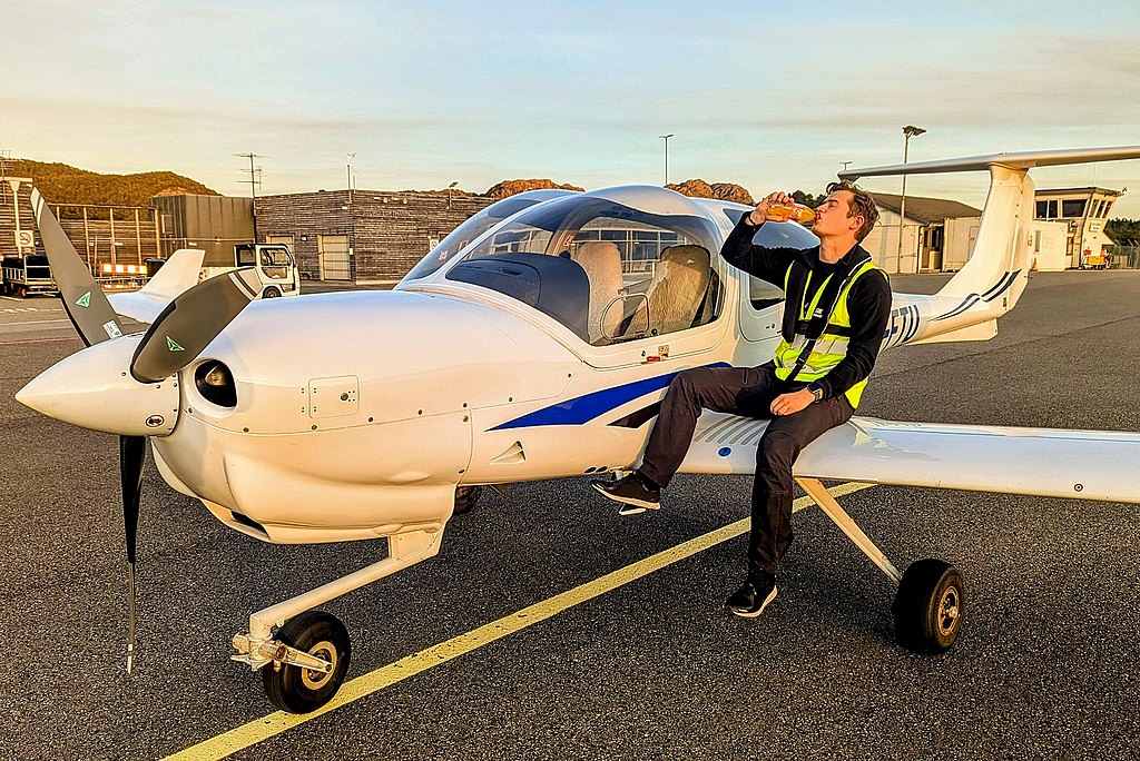 A man in a reflective vest sits on the wing of a DA40 airplane, enjoying a drink against a backdrop of an airport.