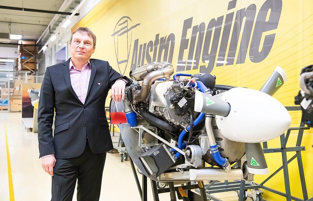A man in a suit stands confidently beside a modern aircraft engine, showcasing advanced engineering in a bright workshop.