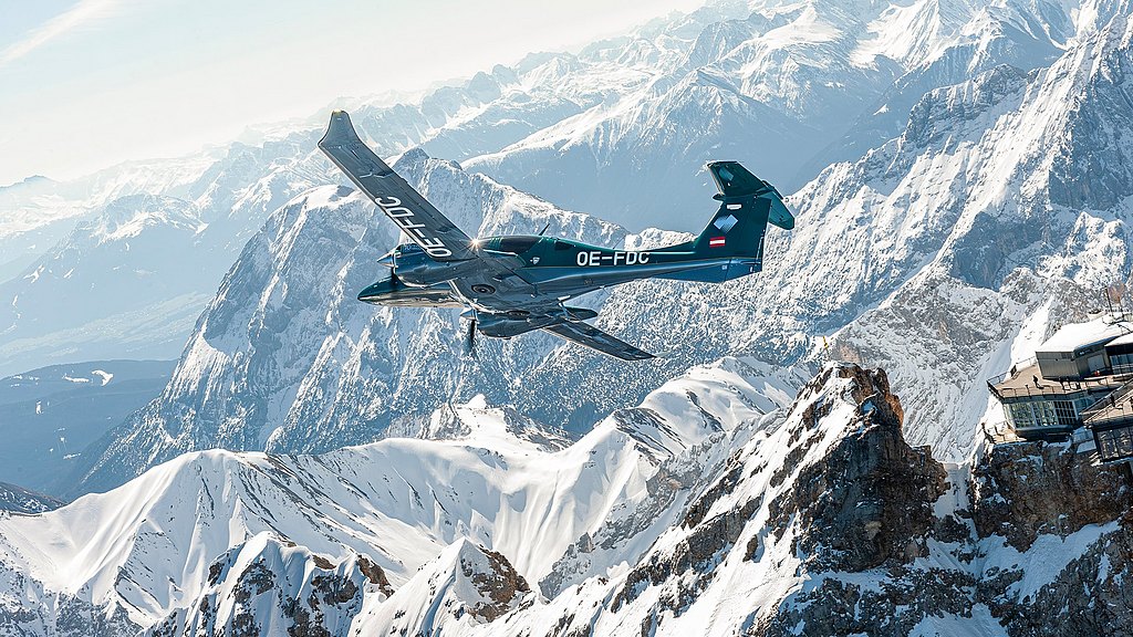 A DA62 aircraft glides gracefully over snow-capped mountains, showcasing a stunning alpine landscape under a clear blue sky.