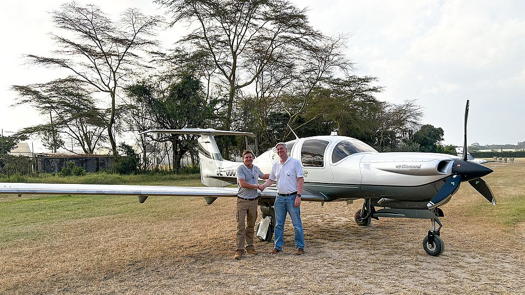 Two men stand beside a sleek DA50 RG, shaking hands in a grassy field surrounded by trees under a cloudy sky.