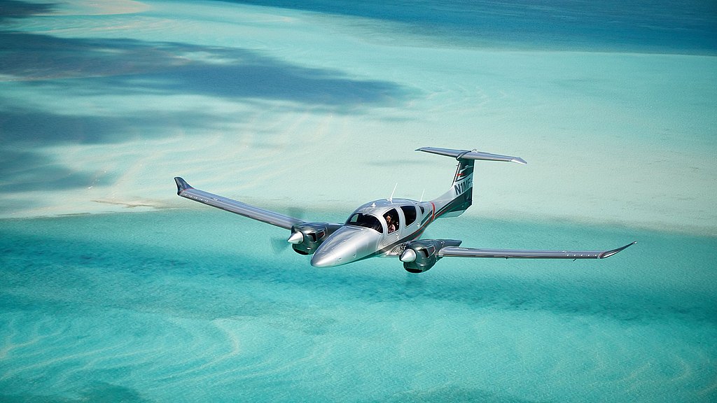 The shiny DA62 plane soars above crystal-clear ocean waters, highlighting the beautiful contrast between the aircraft and the sea below.