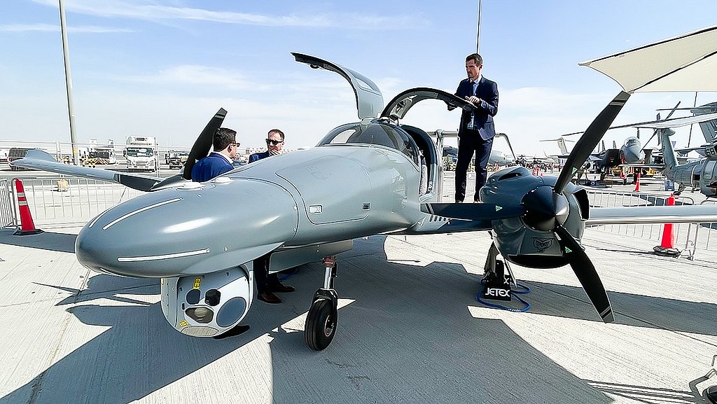 A sleek gray aircraft with its cockpit open, showcasing a man in a suit inspecting the interior while others converse nearby.