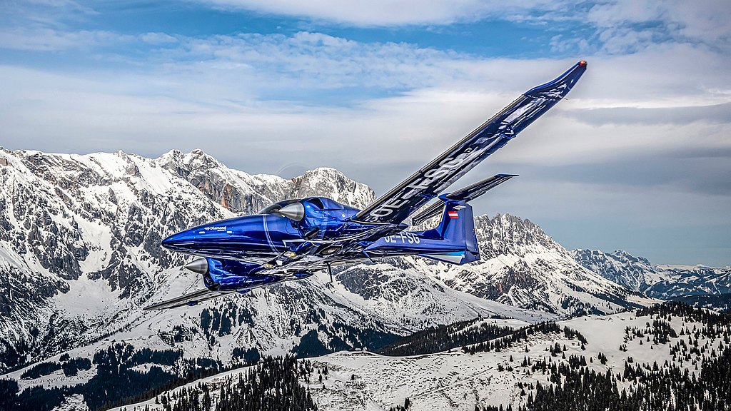 A sleek blue DA62 aircraft soars gracefully above snow-capped mountains, showcasing its aerodynamic design against a clear sky.