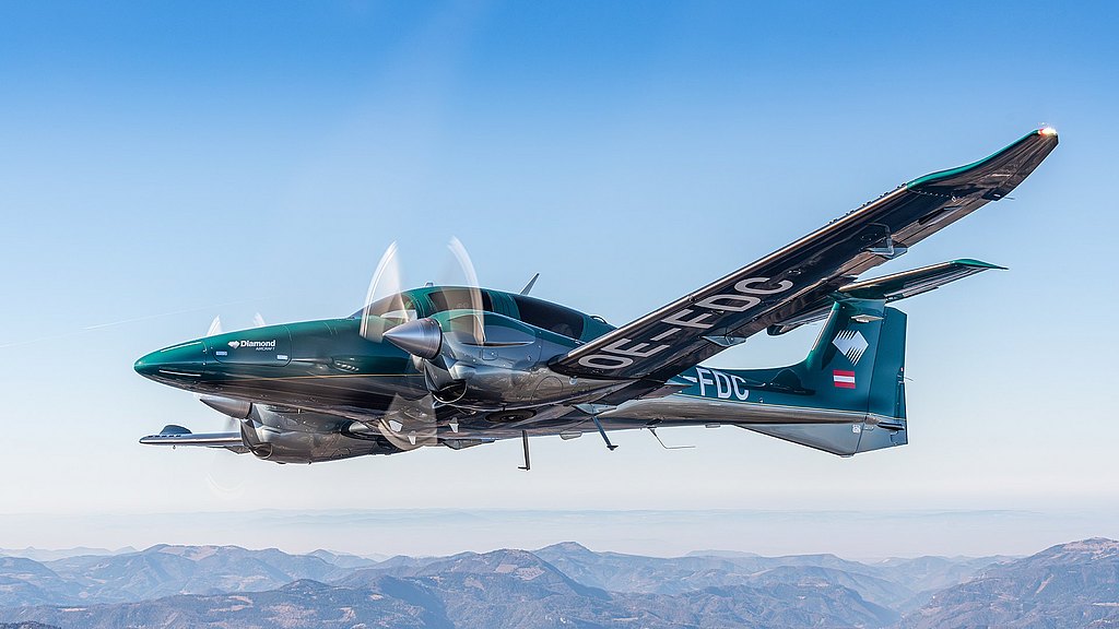 A green DA62 aircraft with spinning propellers soars above a mountainous landscape under a clear blue sky.