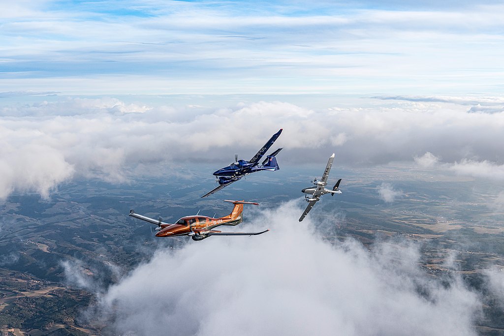 Orange, blue and silver DA62 flying in formation over clouds and landscape.