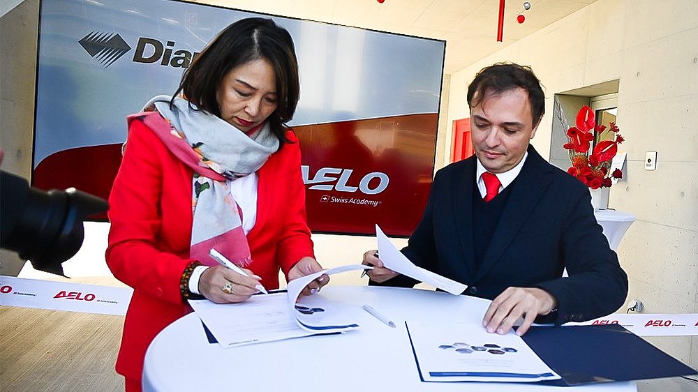 A woman in a red jacket and a man in a black suit are signing documents at a table, surrounded by a modern setting.