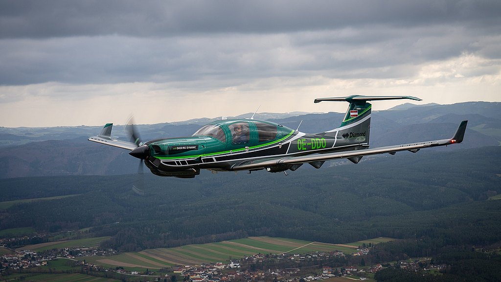 A sleek green and silver DA50 RG aircraft soars through a cloudy sky, showcasing its modern design against a backdrop of rolling hills.