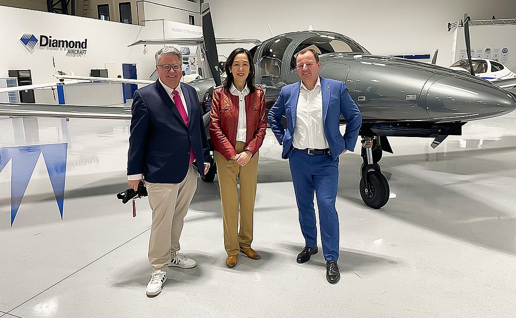 Three professionals stand together in a modern aircraft hangar, smiling in front of a gray DA62 airplane.