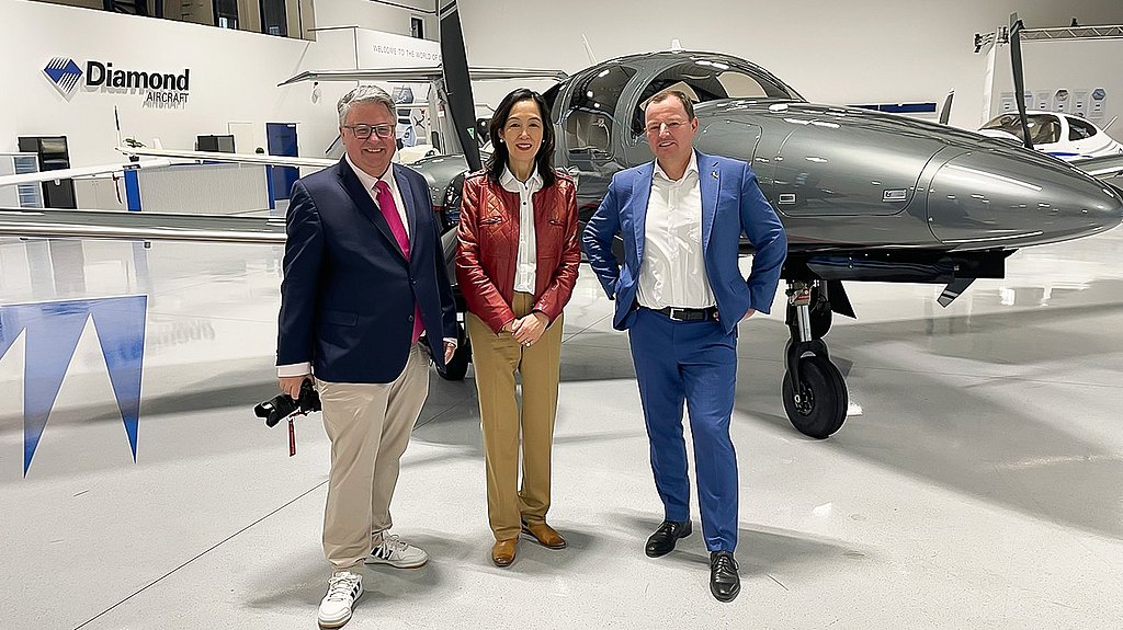 Three professionals stand together in a modern aircraft hangar, smiling in front of a gray DA62 airplane.