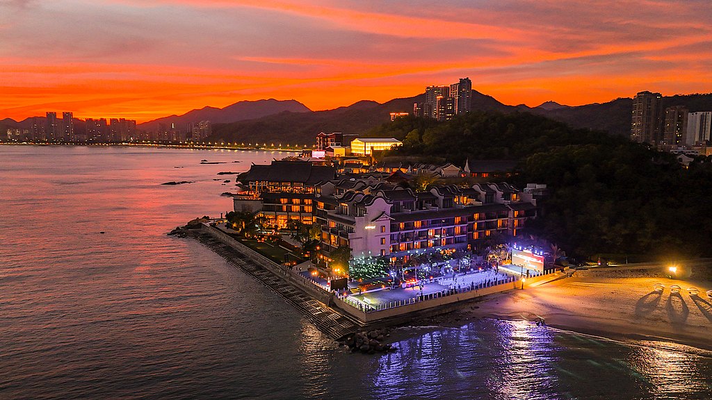 A vibrant sunset casts a warm glow over a coastal resort, highlighting its illuminated buildings and serene beach.