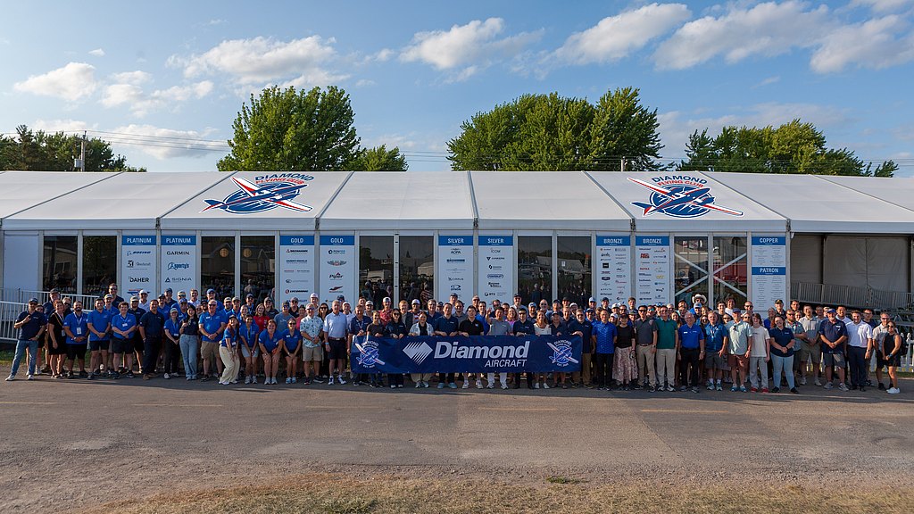 A large group of people stands together in front of a tent, proudly displaying a banner for Diamond Aircraft.