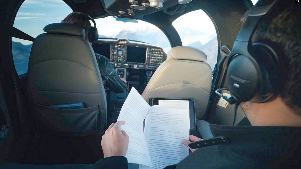 Inside a DA62 aircraft, a passenger reviews documents while the pilot navigates through stunning mountain scenery.