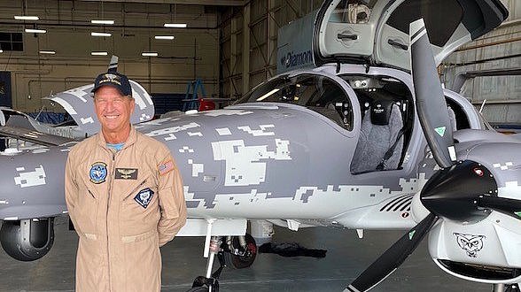 A smiling pilot in a beige flight suit stands proudly next to a gray DA62 MPP aircraft with a unique camouflage pattern.