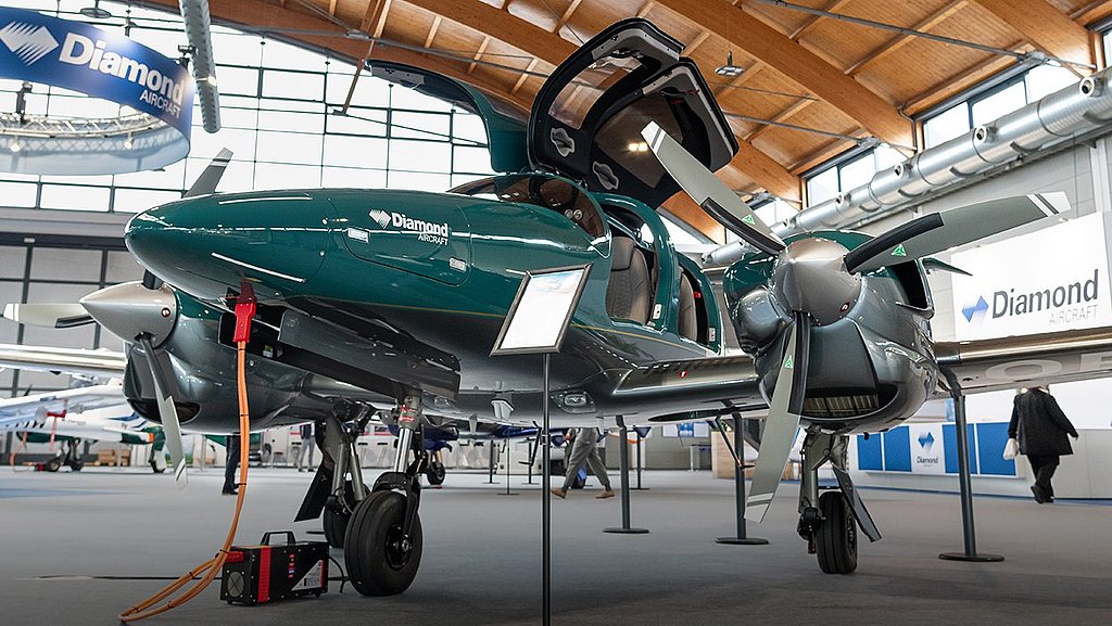 A sleek green DA62 aircraft with its gullwing doors open is showcased at an aviation exhibition, surrounded by other planes and attendees.