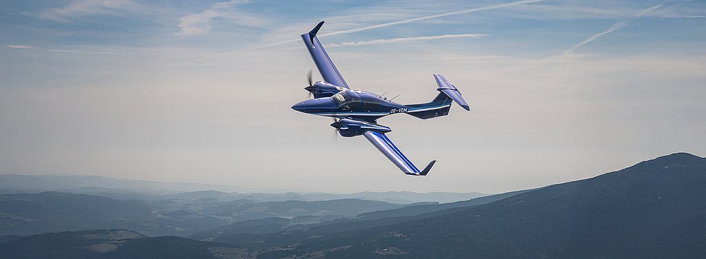 A blue DA42-VI aircraft performs a sharp turn against a backdrop of rolling hills and a clear sky, showcasing its agility.