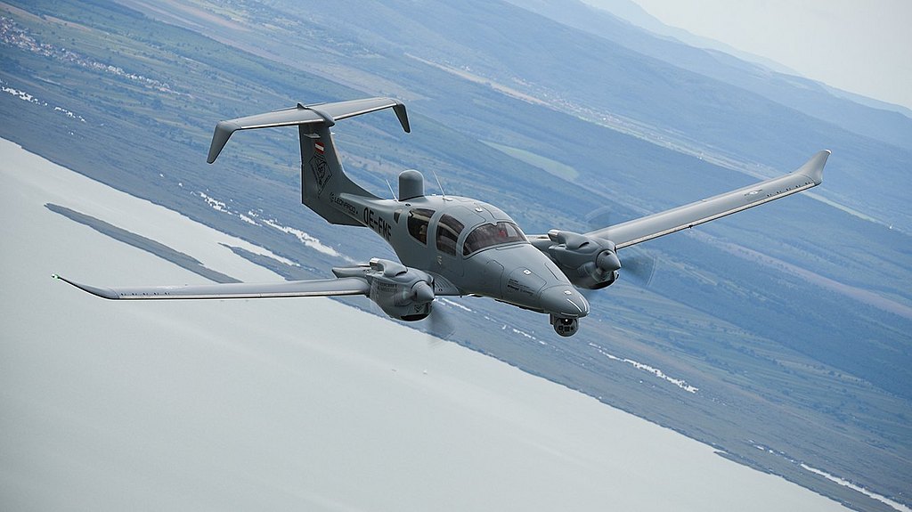 The DA62 MPP aircraft, painted in a muted gray, glides gracefully over a serene body of water, with mountains in the background.