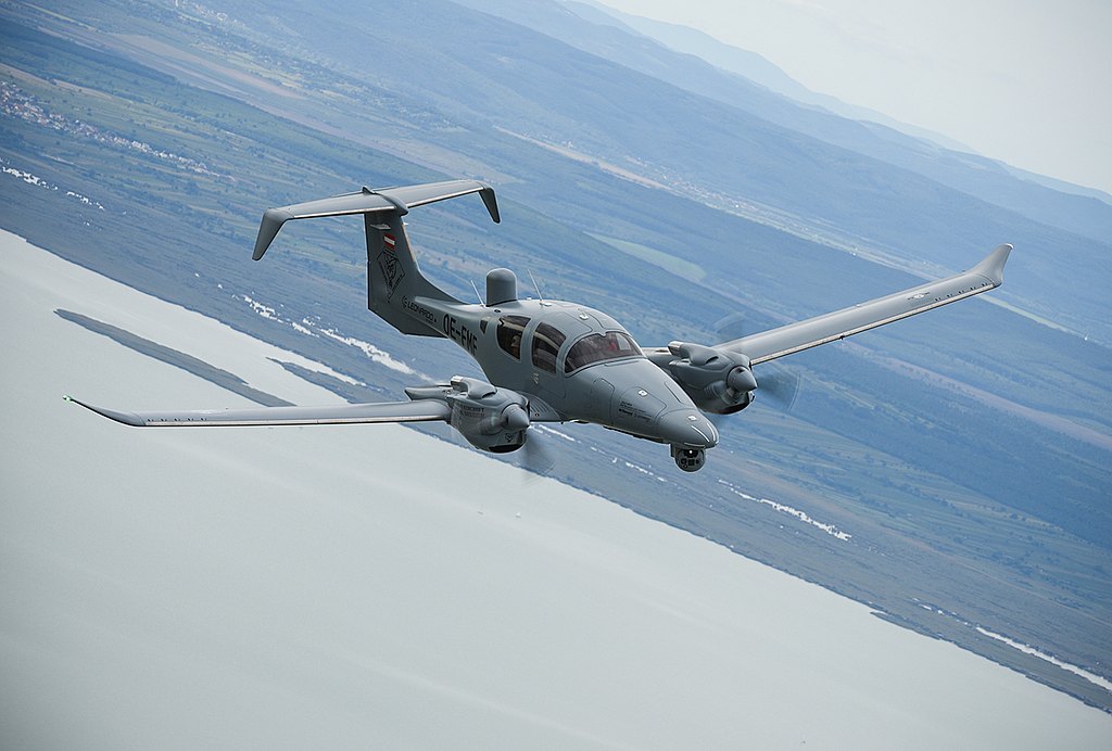 The DA62 MPP aircraft, painted in a muted gray, glides gracefully over a serene body of water, with mountains in the background.