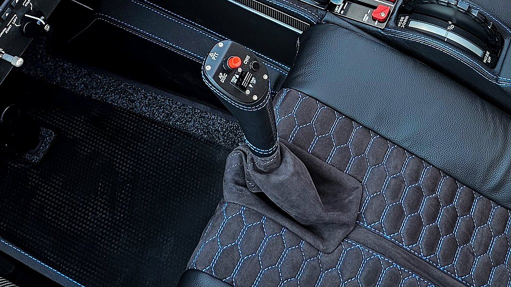 A close-up of a pilot's control stick featuring a red button and sleek black leather with stitching.