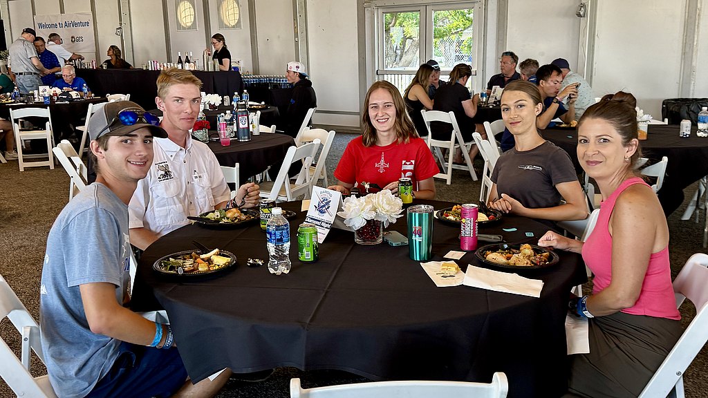 Five individuals sit at a table adorned with flowers, each holding plates of food, creating a warm and friendly atmosphere.
