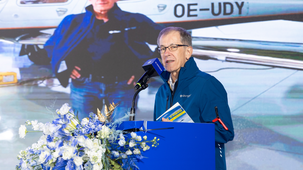 A man in a blue jacket speaks at a podium adorned with flowers, with an airplane image displayed behind him.