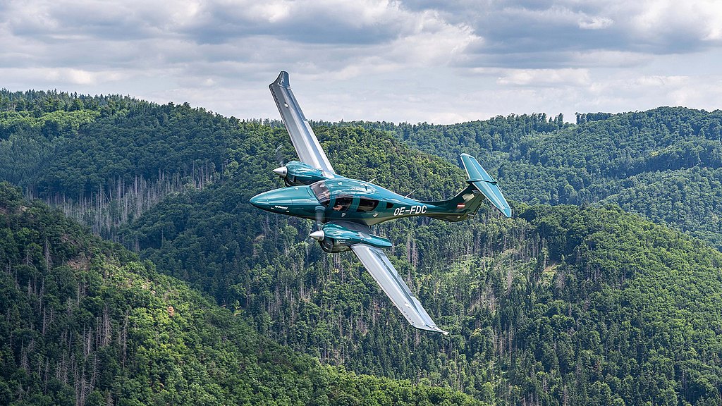 A green DA62 aircraft soars gracefully above lush green hills under a partly cloudy sky, showcasing its elegant design.