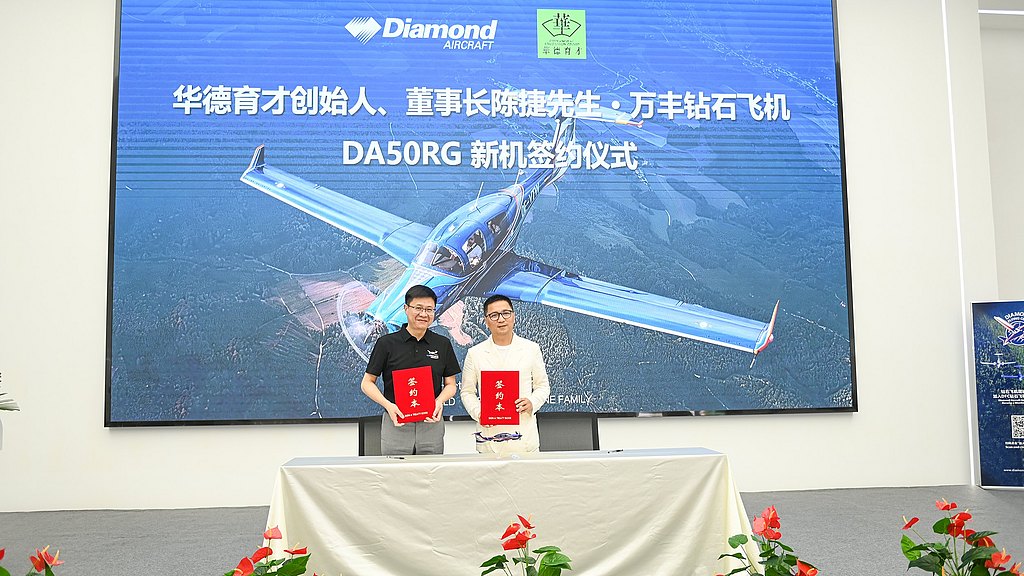 Two men stand at a table, holding red folders, with a large screen displaying an aircraft in the background.