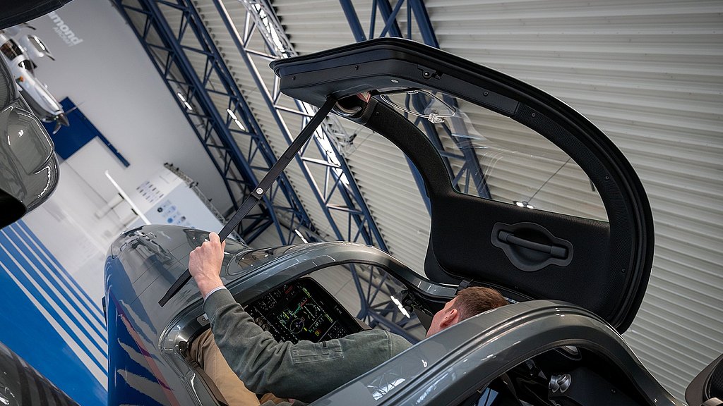 Inside a hangar, an individual grips a door strap, preparing to close the gullwing door of a stylish DA62 aircraft.