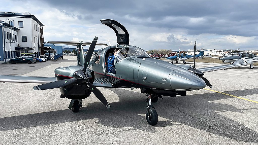 A gray DA62 aircraft with an open gullwing door sits on a tarmac, surrounded by other planes and a cloudy sky.