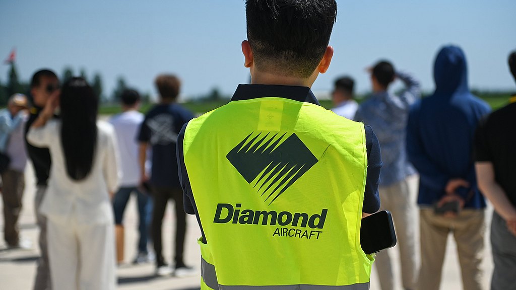 A person in a bright yellow vest with 'Diamond Aircraft' stands among a group, observing an event under a clear blue sky.