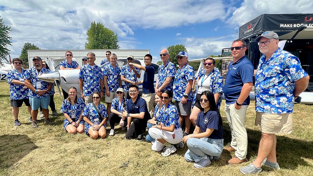 A diverse group of people in matching blue floral shirts poses cheerfully in front of a small airplane on a sunny day.