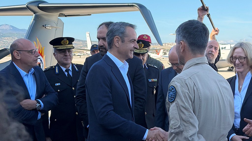 A group of officials, including a man in a pilot's uniform, shake hands warmly at an airport under a clear blue sky.