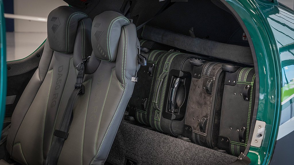 Two modern aircraft seats with green stitching are visible, alongside custom luggage neatly stored behind them.