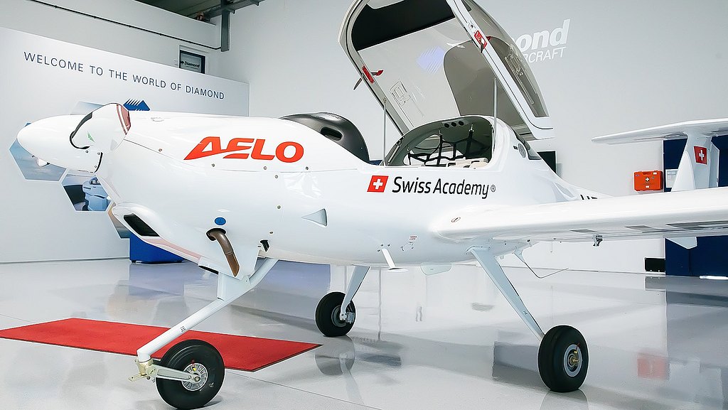 A sleek white DA20i Katana aircraft with 'AELO' branding stands on a polished floor, showcasing its modern design and open cockpit.