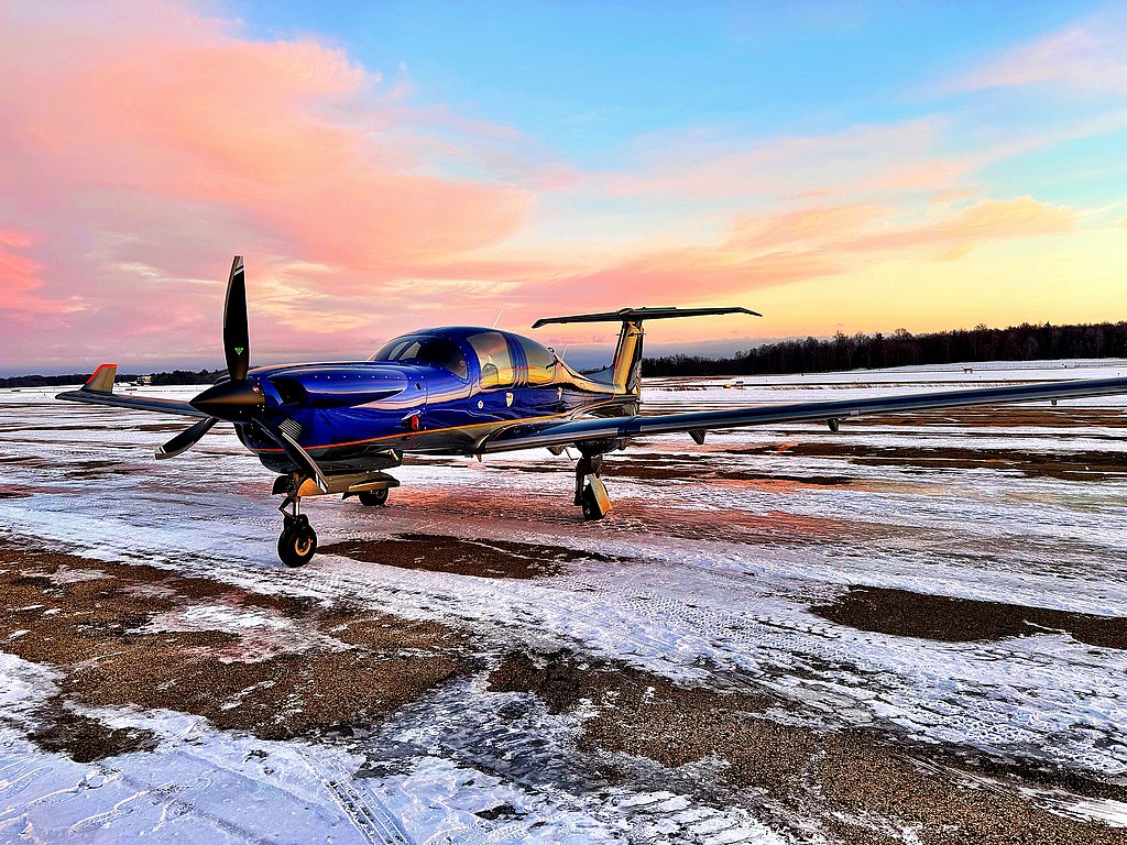 A sleek blue DA50 RG aircraft is parked on a snowy runway, with a vibrant sunset painting the sky in shades of pink and orange.