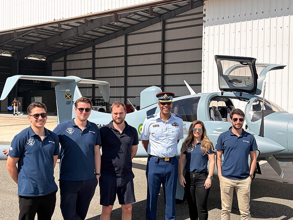 The scene captures a diverse group of people, including a uniformed officer, posing beside a DA62 MPP aircraft in a hangar.