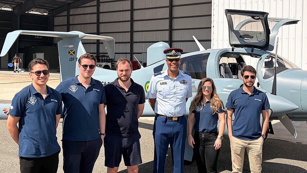 The scene captures a diverse group of people, including a uniformed officer, posing beside a DA62 MPP aircraft in a hangar.