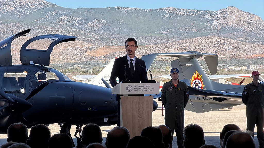 A man in a suit speaks at a podium, flanked by military personnel and an aircraft, with mountains in the background.