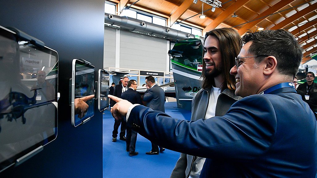 Two men engage enthusiastically with interactive displays at an aviation exhibition, surrounded by attendees and aircraft.